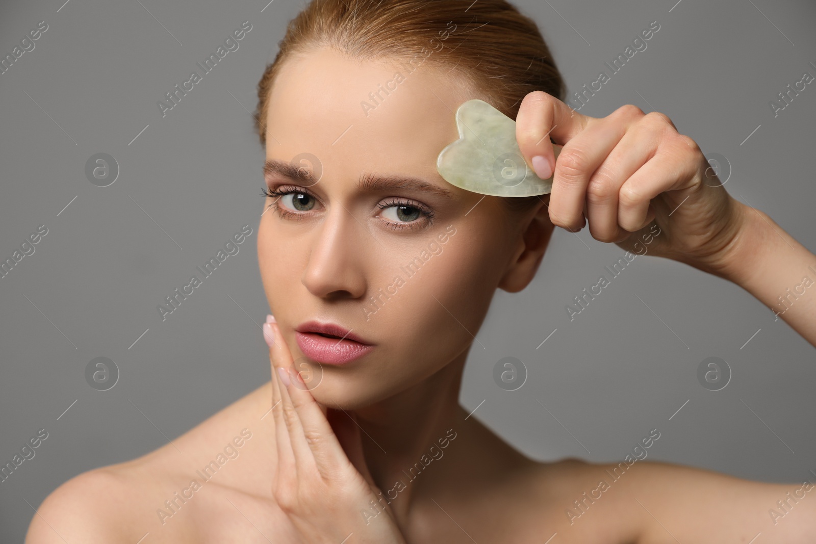 Photo of Beautiful young woman doing facial massage with gua sha tool on grey background, closeup