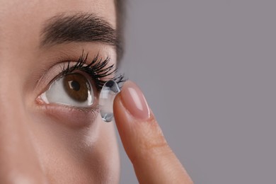 Closeup view of young woman putting contact lens in her eye against grey background. Space for text