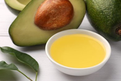 Cooking oil in bowl and fresh avocados on white wooden table, closeup