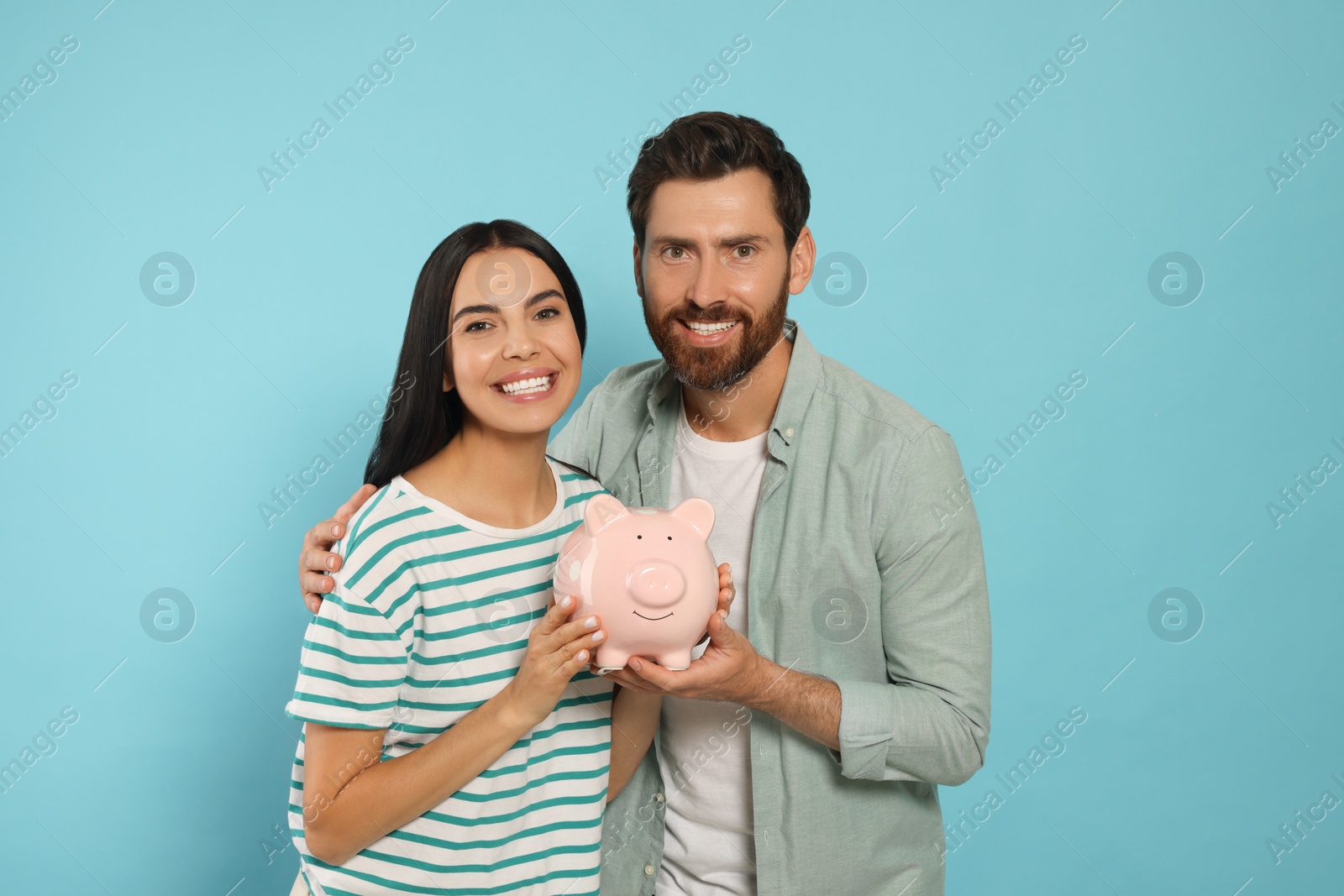 Photo of Happy couple with ceramic piggy bank on light blue background