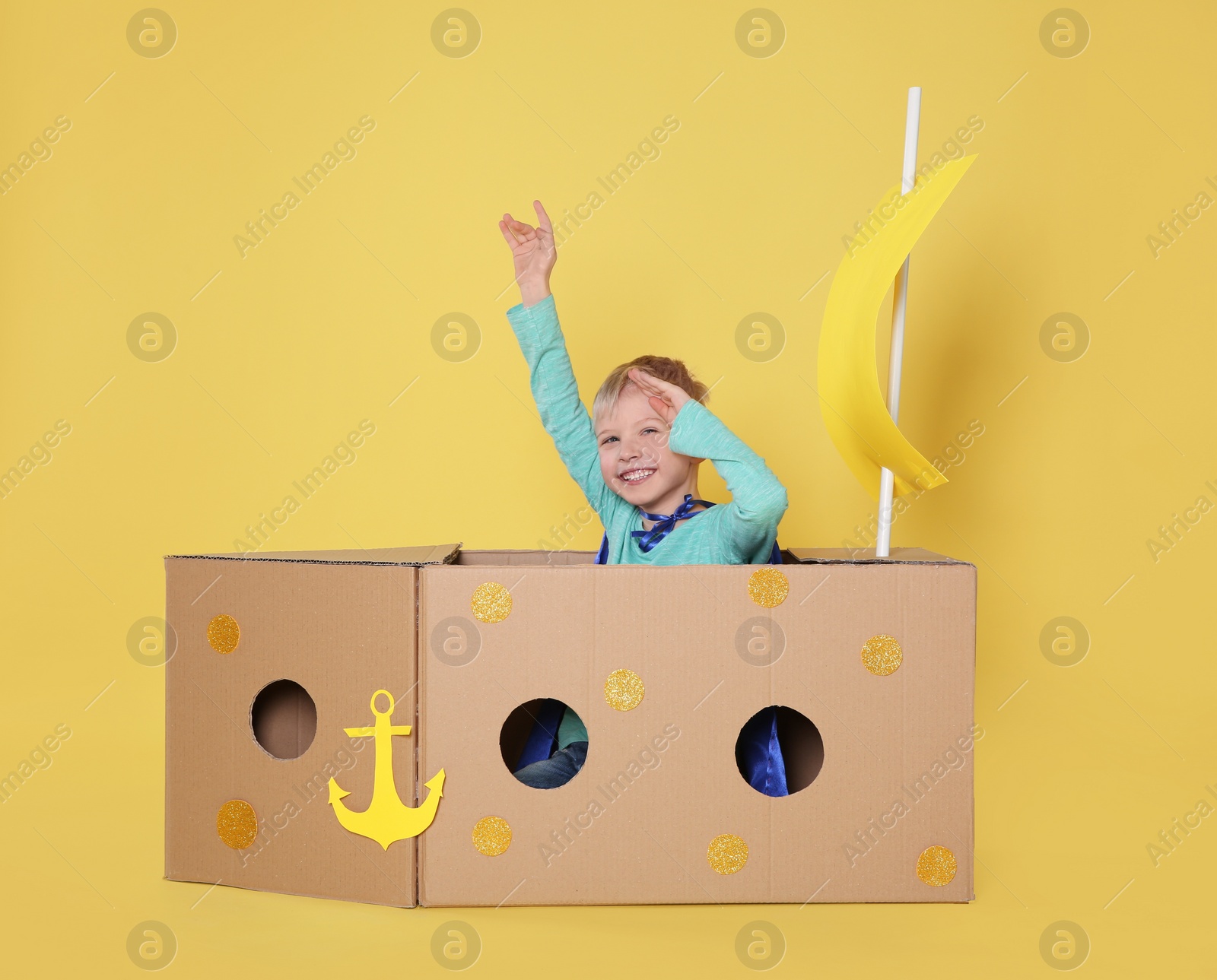 Photo of Little child playing with ship made of cardboard box on yellow background