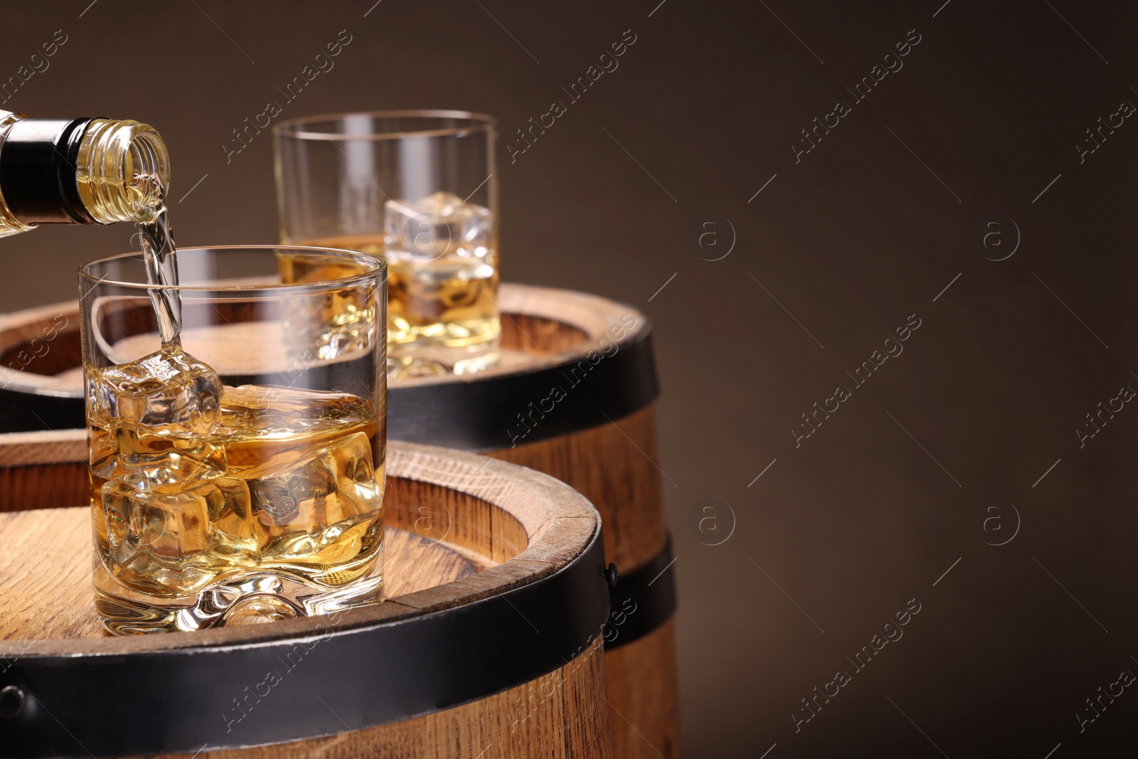 Photo of Pouring whiskey from bottle into glass on wooden barrel against brown background, closeup. Space for text