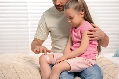 Photo of Father applying ointment onto his daughter's leg on bed indoors
