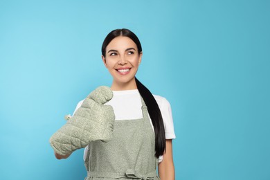 Young woman in grey apron and oven glove on light blue background, space for text