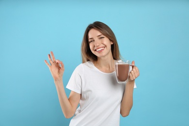 Young woman with glass cup of chocolate milk showing Ok on light blue background