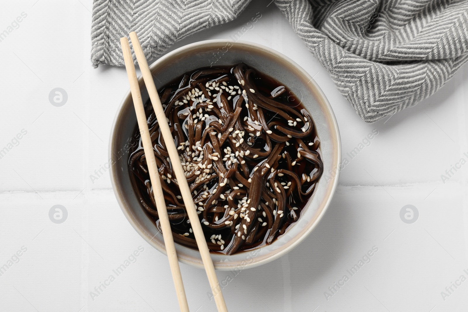 Photo of Tasty buckwheat noodles (soba) with sauce in bowl and chopsticks on white tiled table, top view