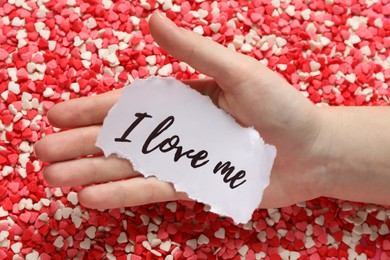 Photo of Woman holding piece of paper with handwritten phrase I Love Me over heart shaped sprinkles, closeup