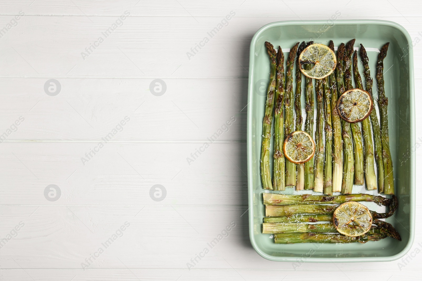 Photo of Oven baked asparagus with lemon slices in ceramic dish on white wooden table, top view. Space for text