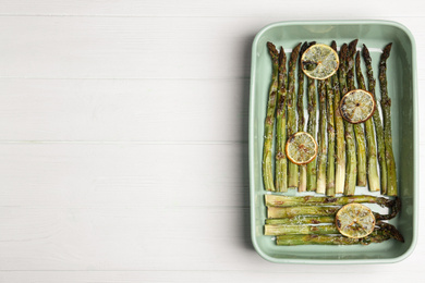 Photo of Oven baked asparagus with lemon slices in ceramic dish on white wooden table, top view. Space for text