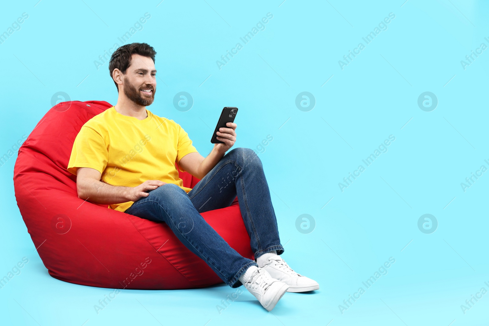 Photo of Smiling man with smartphone sitting on bean bag against light blue background. Space for text