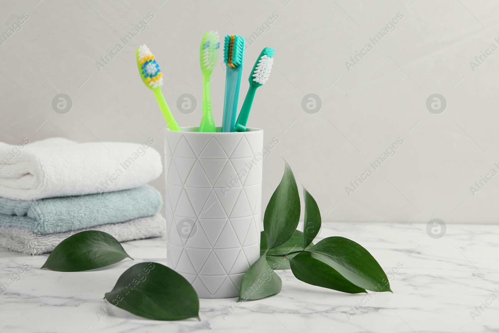 Photo of Colorful plastic toothbrushes in container and green leaves on white marble table, space for text