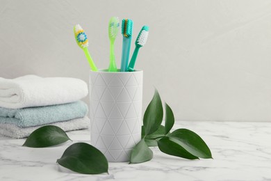 Colorful plastic toothbrushes in container and green leaves on white marble table, space for text