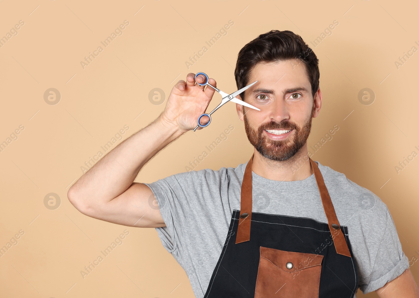 Photo of Smiling hairdresser in apron holding scissors on light brown background