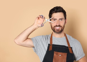 Photo of Smiling hairdresser in apron holding scissors on light brown background