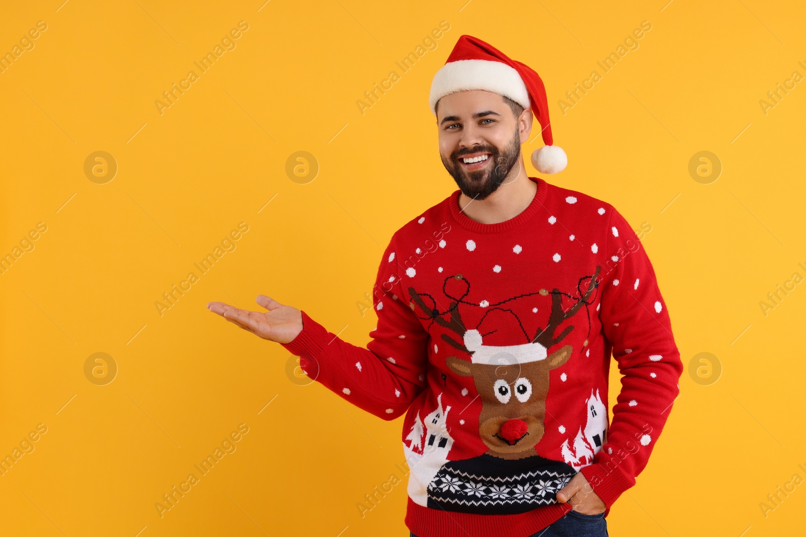 Photo of Happy young man in Christmas sweater and Santa hat showing something on orange background. Space for text