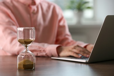 Hourglass with flowing sand on wooden table, selective focus. Man using laptop indoors