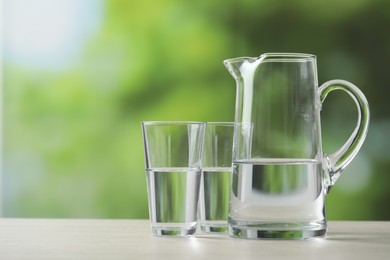 Jug and glasses with clear water on white table against blurred green background. Space for text