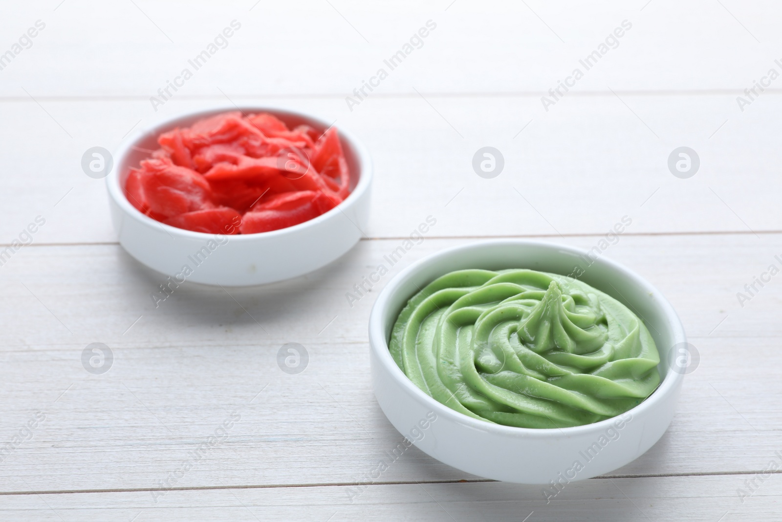 Photo of Bowls with swirl of wasabi paste and pickled ginger on white wooden table, closeup