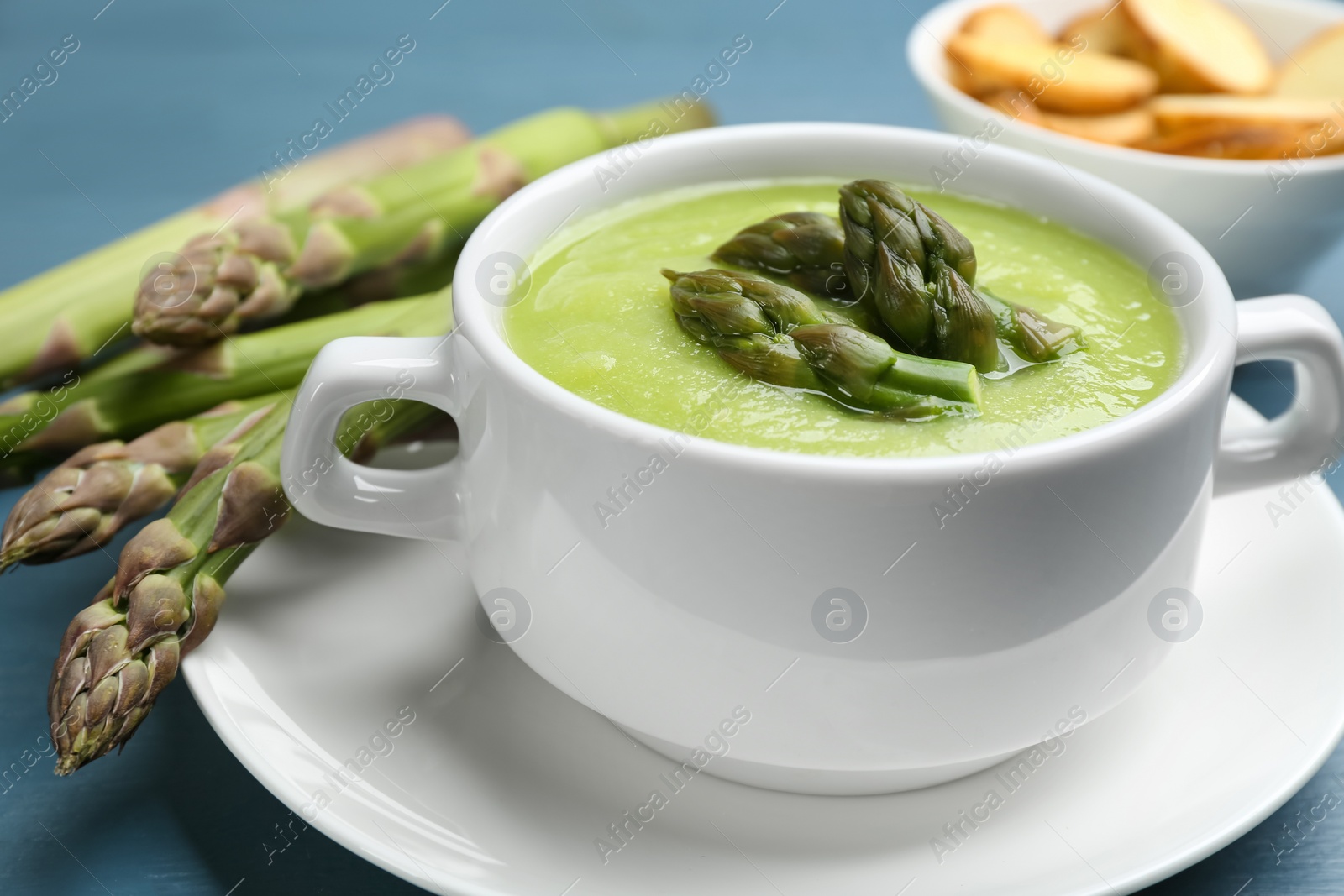 Photo of Delicious asparagus soup served on blue table, 
closeup