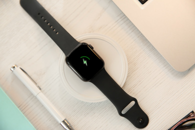 Photo of Flat lay composition with smartwatch and wireless charger on white wooden table. Modern workplace accessory