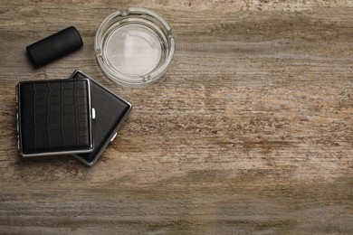 Closed cigarette cases, lighter and empty glass ashtray on wooden table, flat lay. Space for text