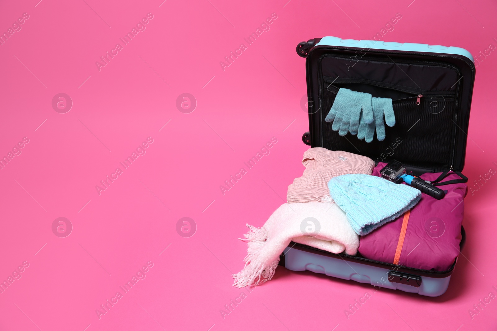 Photo of Open suitcase with warm clothes and action camera on color background, space for text. Winter vacation