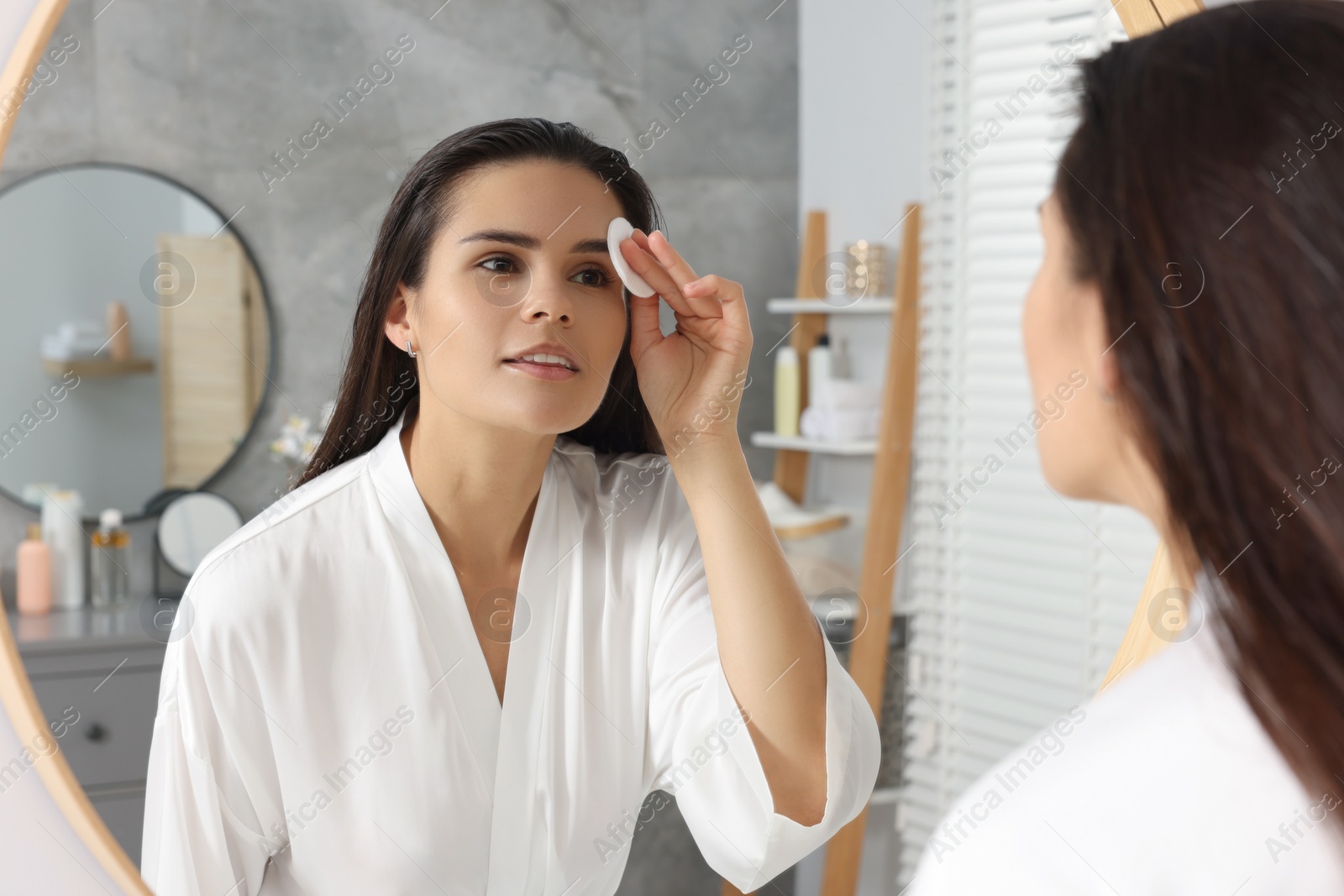 Photo of Young woman cleaning her face with cotton pad near mirror in bathroom