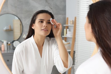 Young woman cleaning her face with cotton pad near mirror in bathroom