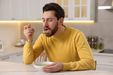Man eating delicious chicken soup at light marble table in kitchen