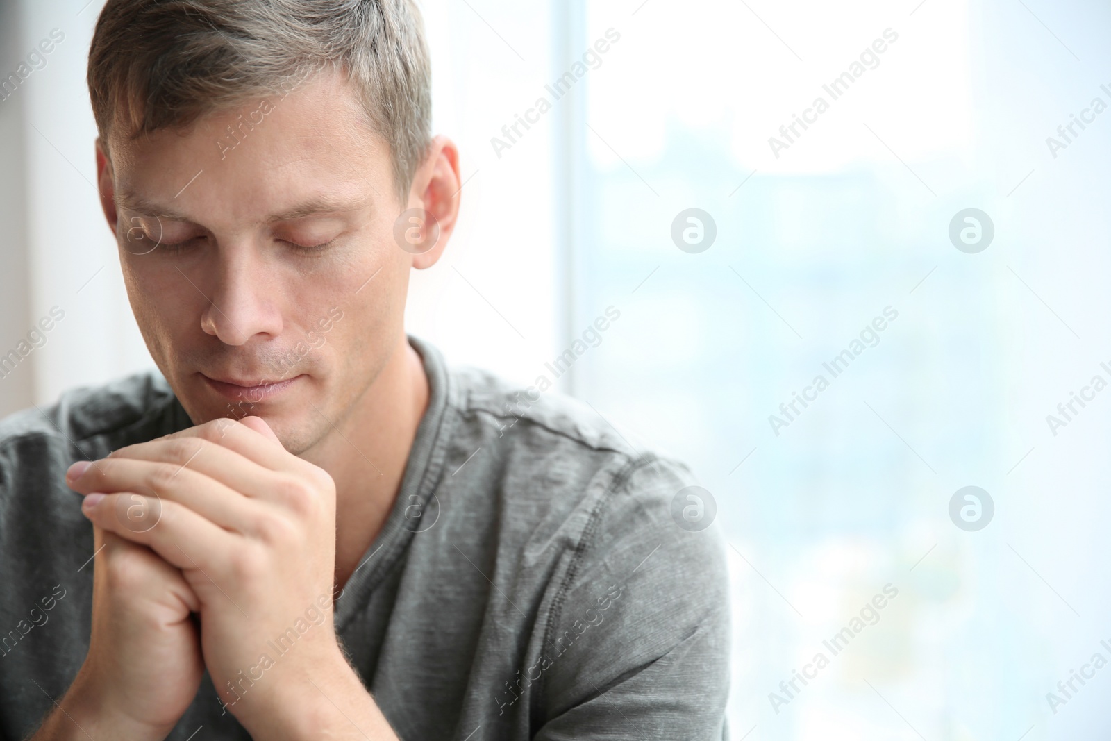 Photo of Man with hands clasped together for prayer near window. Space for text