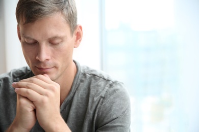 Photo of Man with hands clasped together for prayer near window. Space for text