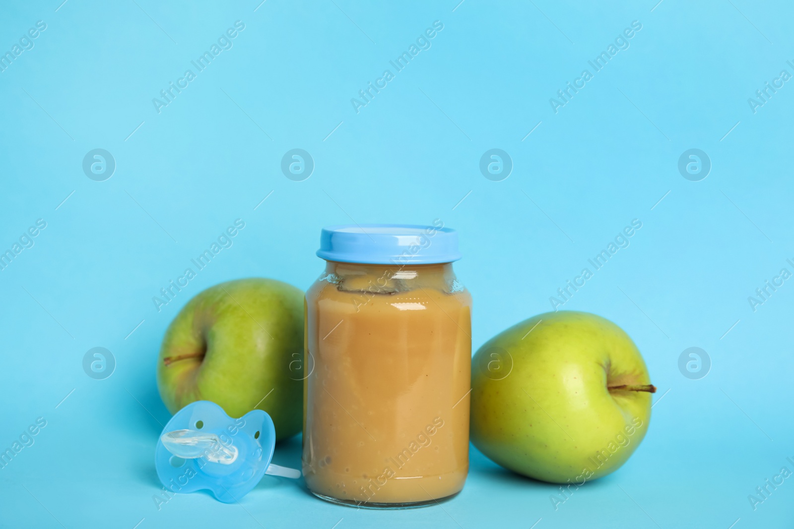 Photo of Baby food in jar, pacifier and fresh apples on light blue background