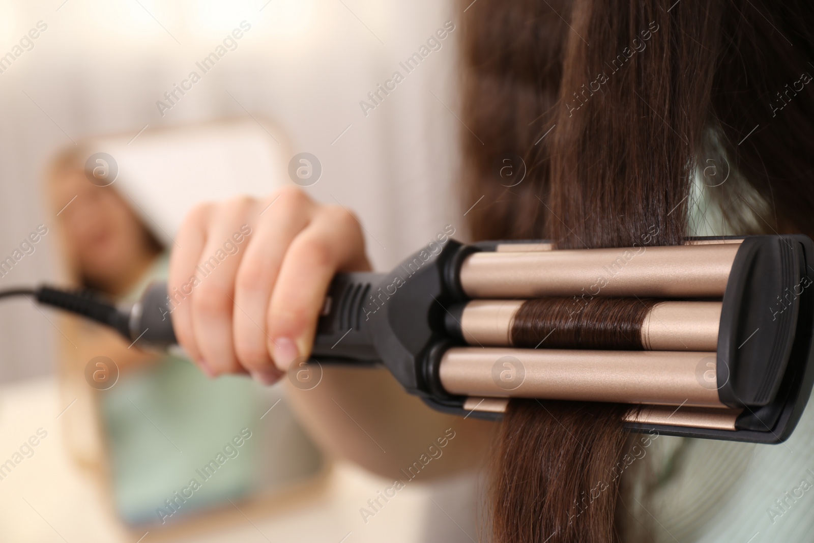 Photo of Young woman using modern curling iron indoors, closeup