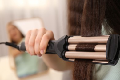 Photo of Young woman using modern curling iron indoors, closeup