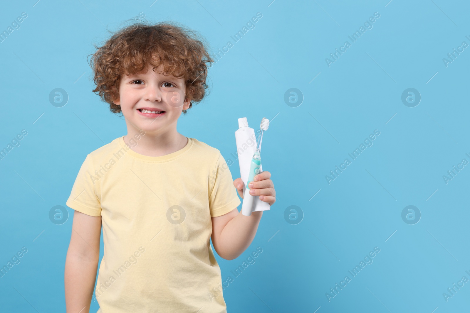 Photo of Cute little boy holding electric toothbrush and tube of toothpaste on light blue background, space for text