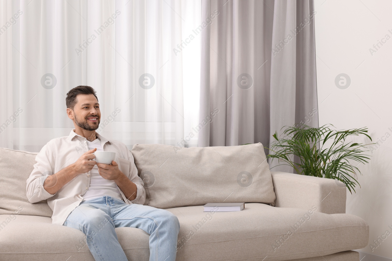 Photo of Happy man drinking coffee while resting on sofa near window with beautiful curtains in living room. Space for text