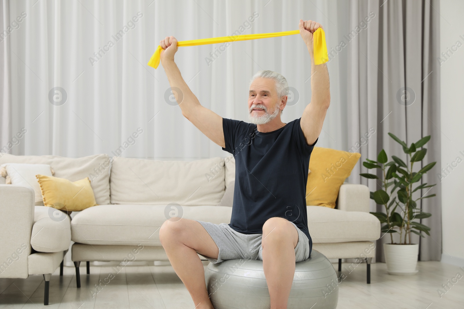 Photo of Senior man doing exercise with elastic resistance band on fitness ball at home