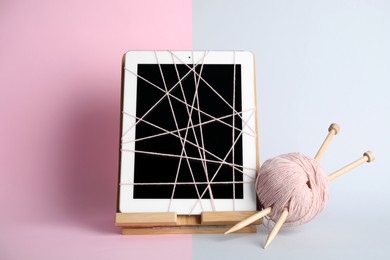 Modern tablet wrapped in thread, yarn ball and knitting needles on color background. Internet addiction concept