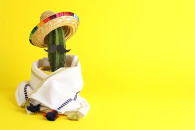 Photo of Cactus with Mexican sombrero hat, fake mustache and poncho on yellow background, space for text