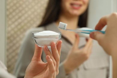 Young woman with toothbrush and bowl of baking soda near mirror indoors, closeup