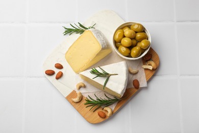 Photo of Pieces of tasty camembert cheese, rosemary, nuts and olives on white tiled table, top view
