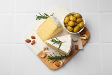 Pieces of tasty camembert cheese, rosemary, nuts and olives on white tiled table, top view