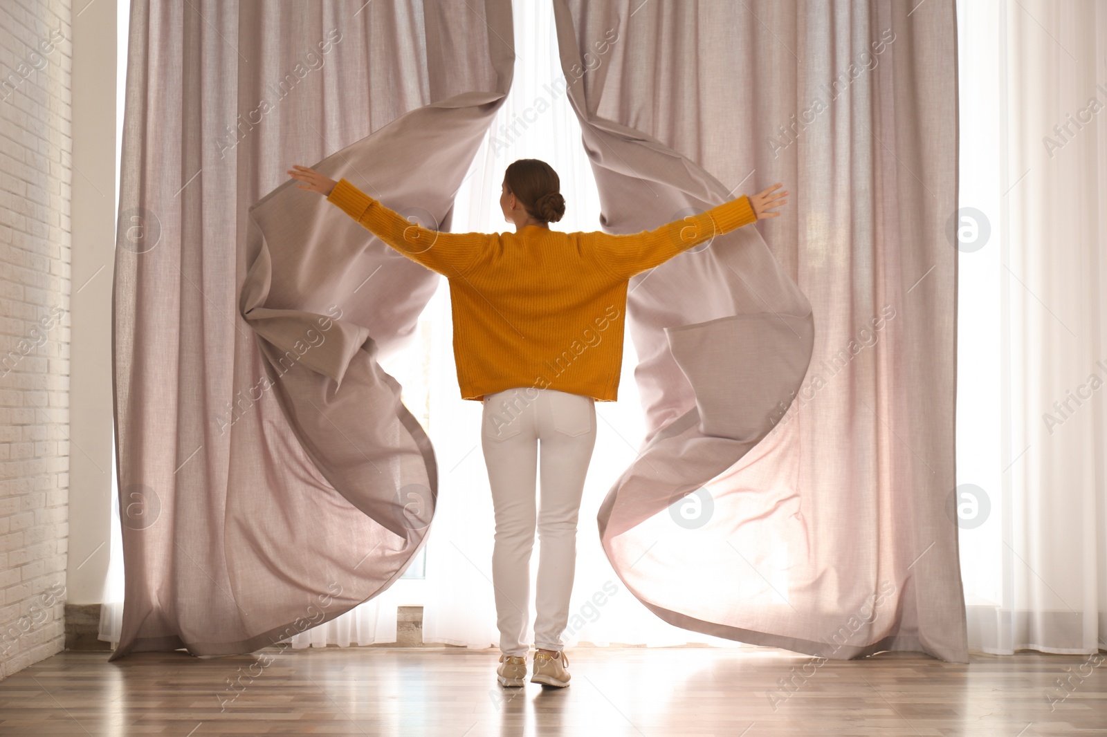 Photo of Woman opening window curtains at home in morning, back view