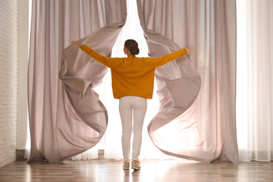 Photo of Woman opening window curtains at home in morning, back view