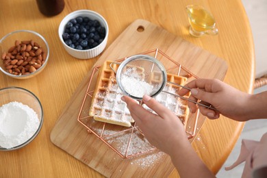 Woman decorating delicious Belgian waffles with powdered sugar at wooden table, closeup