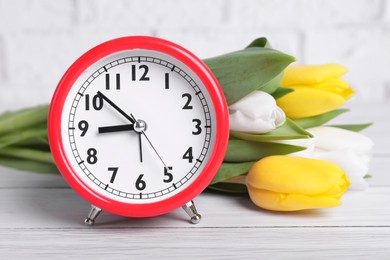 Photo of Red alarm clock and beautiful tulips on white wooden table against brick wall, closeup. Spring time