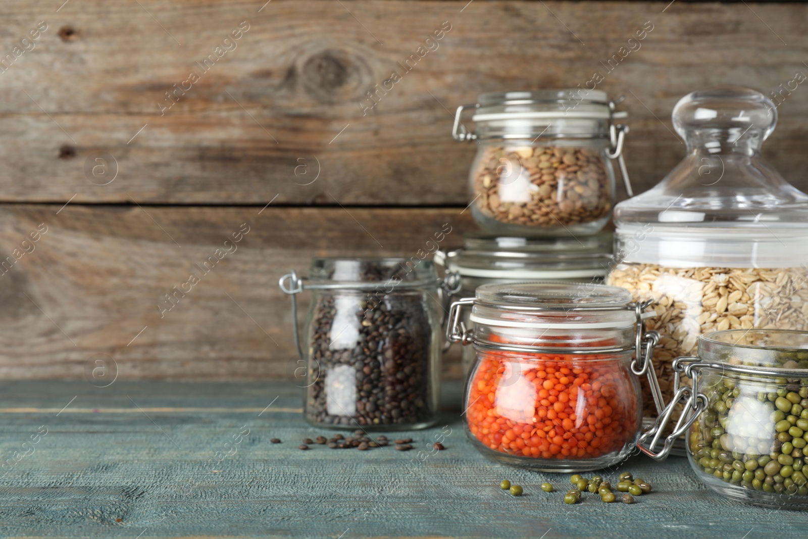 Photo of Different types of legumes and cereals in glass jars on blue wooden table, space for text. Organic grains