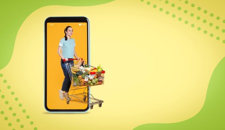 Image of Happy woman with shopping cart full of groceries walking out huge smartphone on color background. Design with space for text