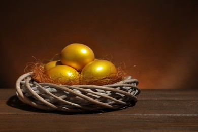 Photo of Shiny golden eggs in nest on wooden table, space for text