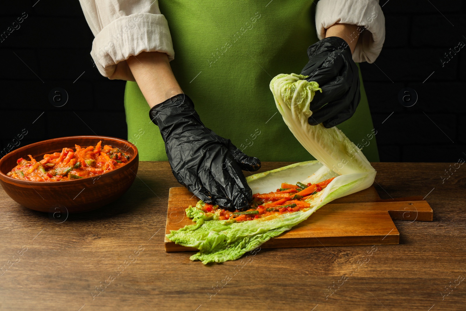 Photo of Woman preparing spicy cabbage kimchi at wooden table against dark background, closeup